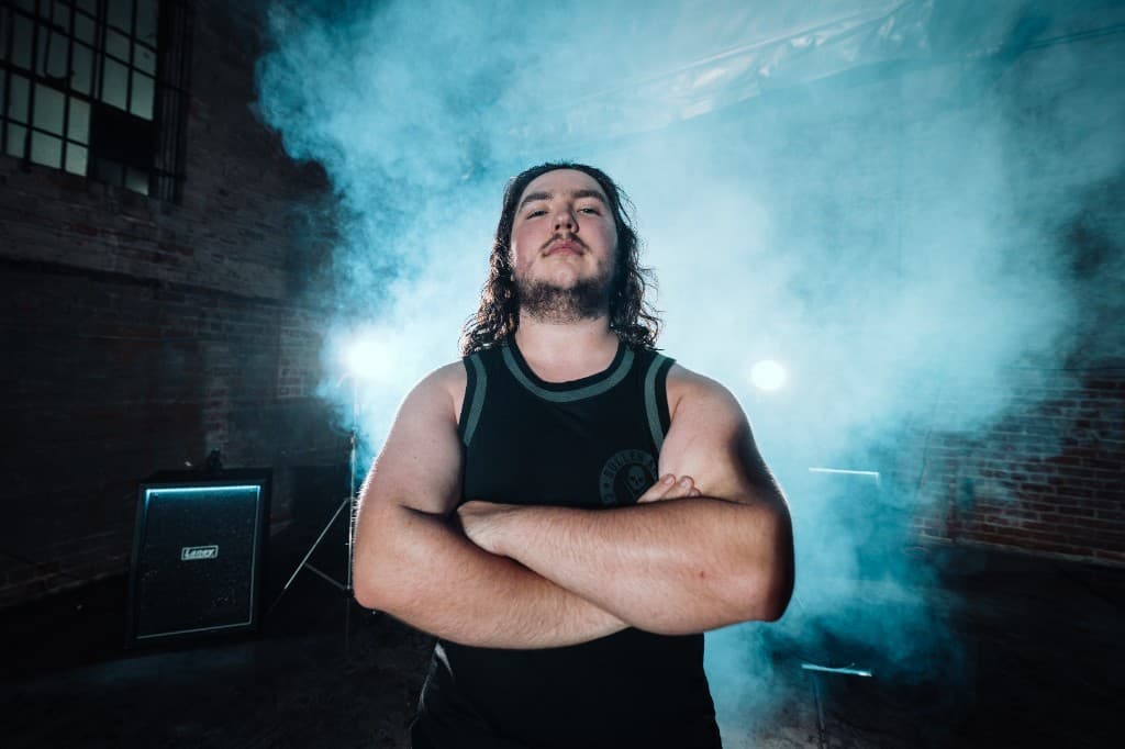 Maxx of Fall of Damascus, arms crossed in front of a wall of smoke